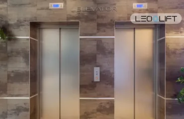 Two stainless-steel elevator doors in a marble-tiled wall, with a central call button panel and potted plants on both sides.