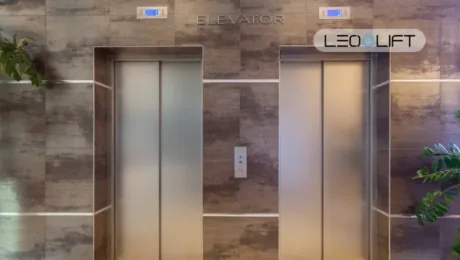 Two stainless-steel elevator doors in a marble-tiled wall, with a central call button panel and potted plants on both sides.