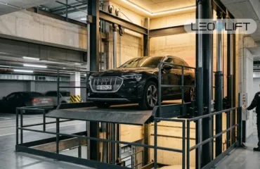 Avtomobilnyy lift dlya parkinhu koly vin vyhidnishyy za rampu Black Audi on a car elevator platform in a dim underground parking garage, with a technician nearby.