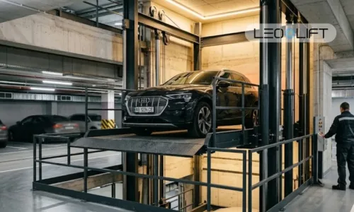 Black Audi on a car elevator platform in a dim underground parking garage, with a technician nearby.