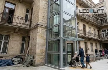 Dostupnist pislya rekonstruktsiyi yak dodaty lift u staru budivlyu Entrance to a stone building with a tall glass elevator shaft; a man pushes an elderly person in a wheelchair, a child follows, and a group of people waits nearby under the LEOLIFT logo.