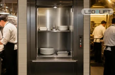 Stainless steel pass-through lift in a busy commercial kitchen, with plates and bowls neatly stacked on its shelves.