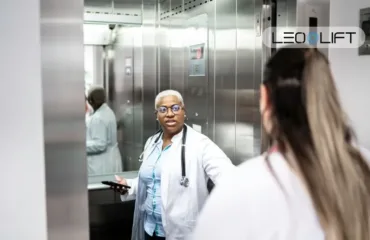 Likarnyanyy lift rozmiry kabiny, dveri, nosylky ta bezpechne perevezennya African American female doctor in a white coat with a stethoscope, holding a phone, conversing in a hospital elevator with a colleague nearby.