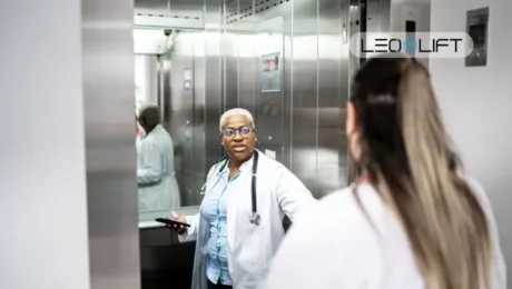 African American female doctor in a white coat with a stethoscope, holding a phone, conversing in a hospital elevator with a colleague nearby.