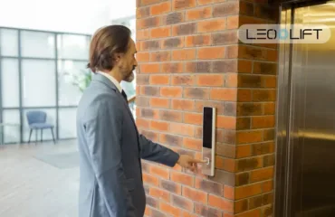 Man in a gray suit presses the elevator call button mounted on a brick wall, with a LeoLift logo nearby.