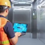 Maintenance technician in an orange safety vest and hard hat uses a laptop to monitor a modern elevator system in a metallic shaft area.