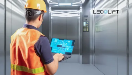 Maintenance technician in an orange safety vest and hard hat uses a laptop to monitor a modern elevator system in a metallic shaft area.