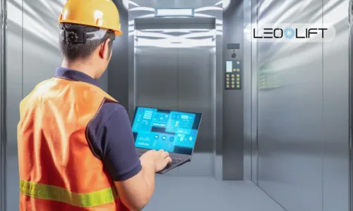 Maintenance technician in an orange safety vest and hard hat uses a laptop to monitor a modern elevator system in a metallic shaft area.