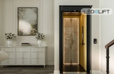 Domashniy lift bez mashynnoho prymishchennya koly tse naykrashche rishennya Modern home entrance with a lift/elevator door opened to a gold-toned interior, flanked by a white console table and framed wall art